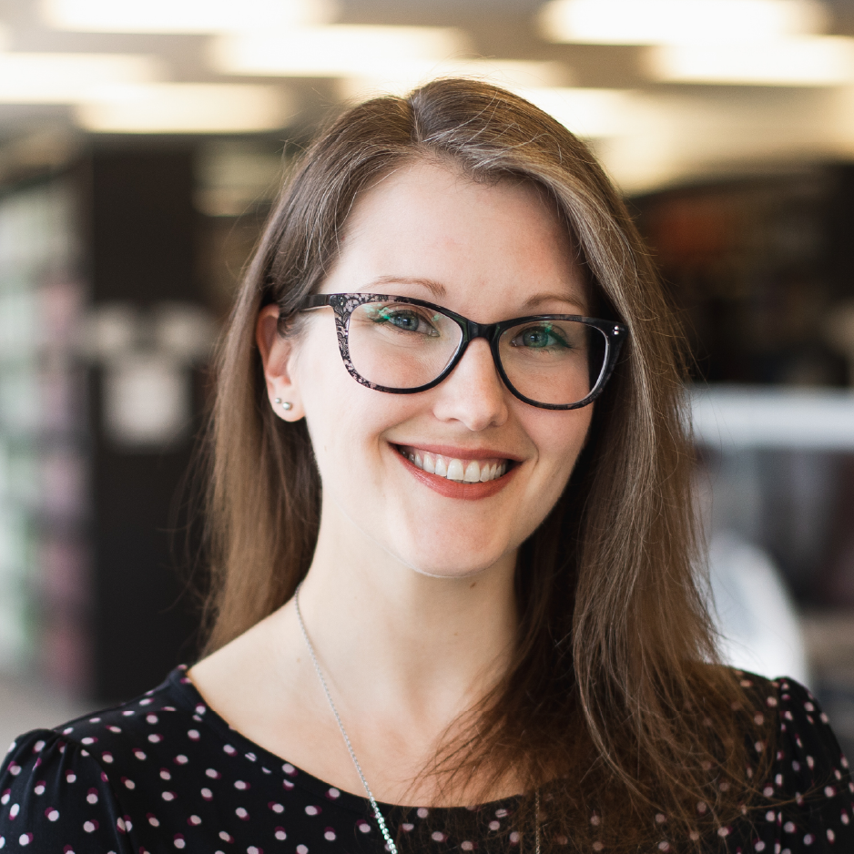 Stephanie Mutch with long brown hair and black glasses wearing a black blouse with light coloured dots standing in an indoor setting with blurred background