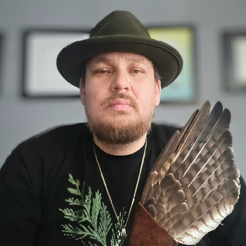 Kevin George wearing a black hat and black shirt with a cedar motif holding an eagle feather in an indoor setting with blurred background