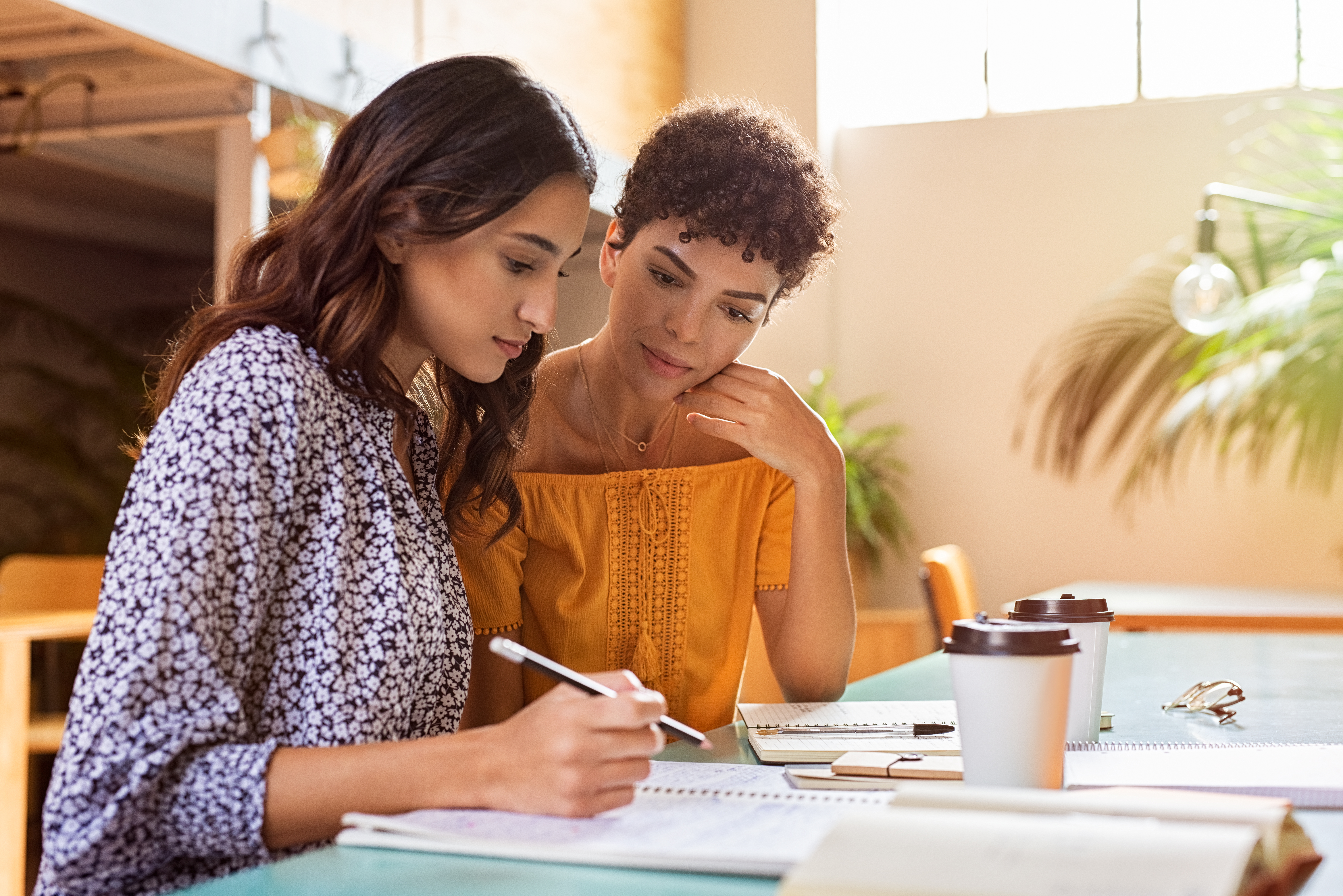 Young multiethnic women studying together at library. High school or college students studying and reading together in cafeteria. Latin girl explaining to brazilian friend the lesson.