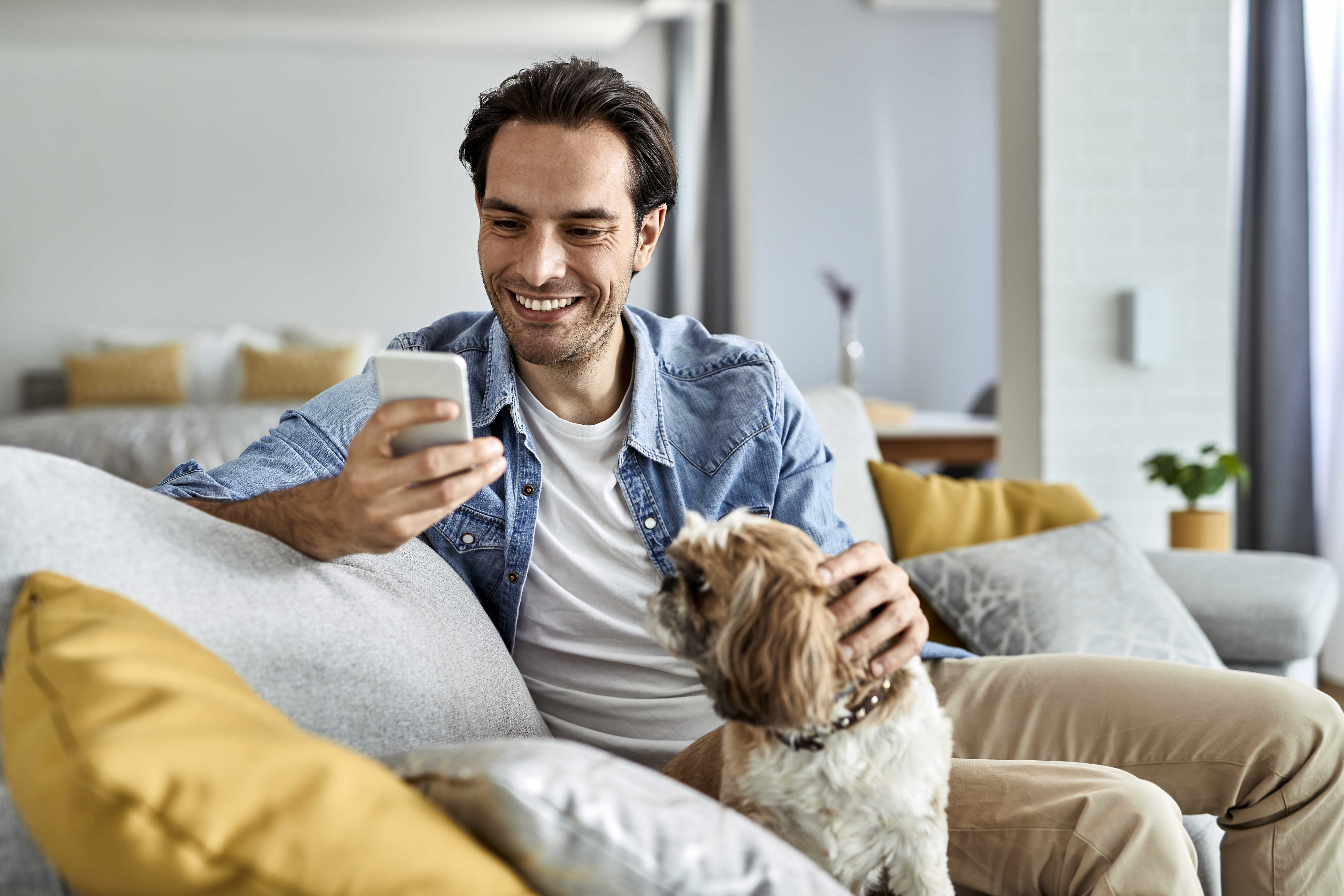 Young happy man text messaging on cell phone while sitting on the sofa with his dog.