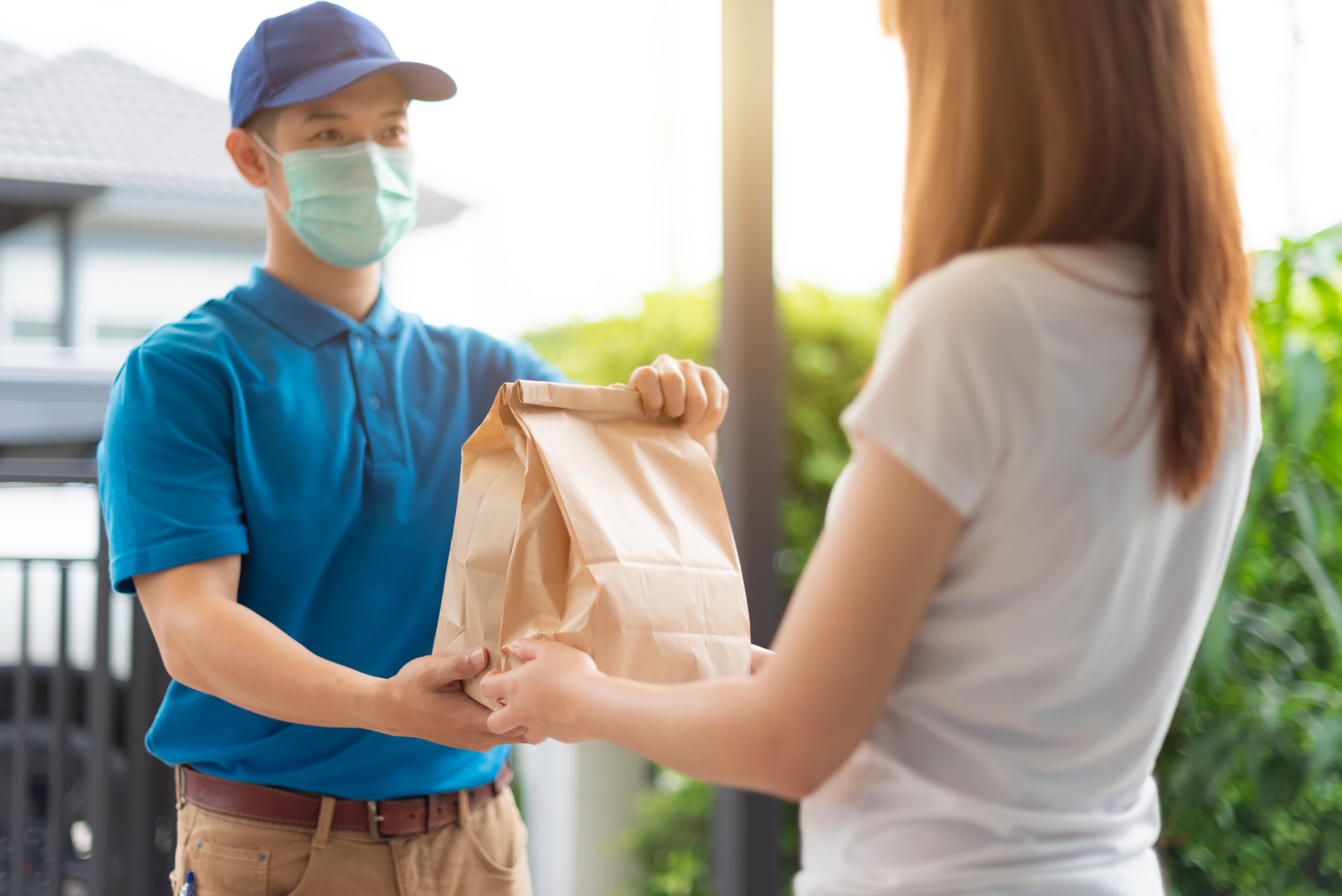 The delivery man is delivering the paper bag to beautiful female customer in front of the home. The delivery man in a blue dress delivered a bag of food to the customer. online delivery concept.