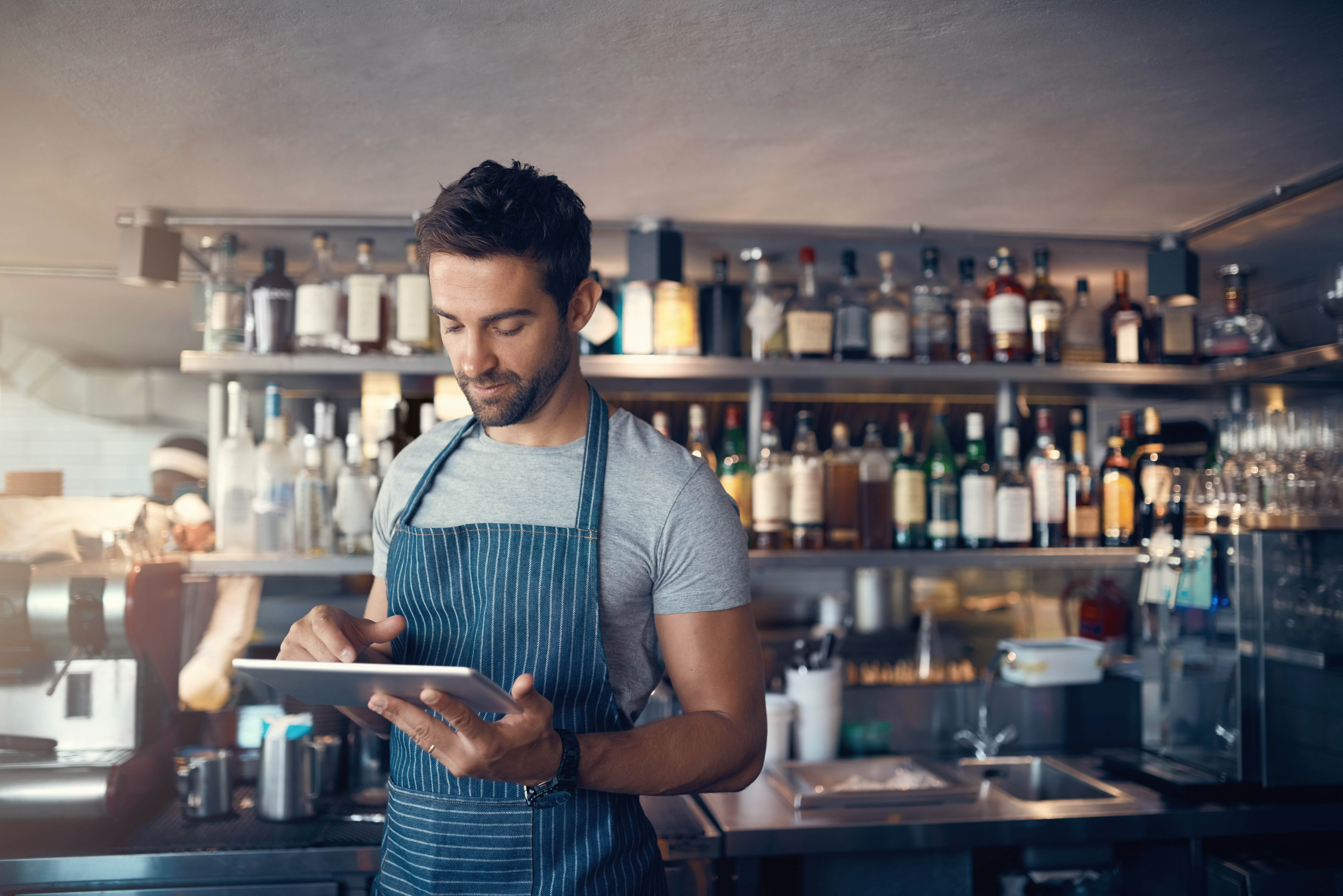 man with apron in front of shelves of alcohol in bar of restaurant looking down at tablet in his hands